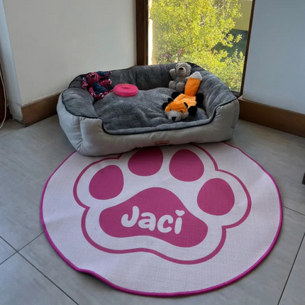 A gray plush pet bed with stuffed toys sits on a light tile floor next to a large round white rug featuring a pink paw print and the name 'Jaci' in white. A window in the background shows green trees and outdoor scenery.