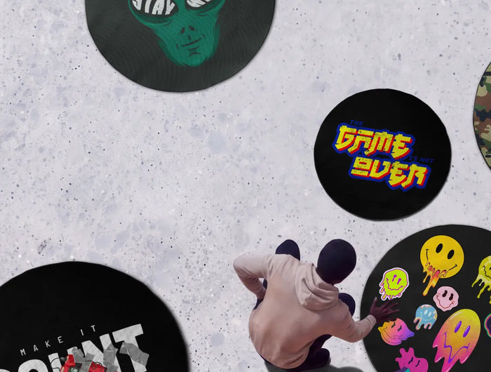 Promotional banner featuring a light concrete-textured background with several large circular rugs in colorful graphic designs, including lips, an alien, camouflage, retro gaming, smiley faces, an astronaut, and cartoon artwork. A blue badge in the top-left corner advertises 30% OFF with the code 30OFF. In the top-right corner, a person in a white hoodie and sweatpants relaxes on a large blue bean bag while reading a book. A person is also shown interacting with one of the rugs near the bottom center.